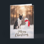 Boston Terrier Christmas Snowman Feestdagen Kaart<br><div class="desc">A charming winter holiday scene featuring a Boston Terrier sitting in fresh snow beside a cheerful little snowman wrapped in a bright red scarf. Soft golden winter light and gently falling snow create a peaceful seasonal moment that highlights the expressive face and playful personality Boston Terriers are known for. Perfect...</div>