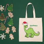 Cute Kerstmis Dinosaur Custom Green Kinder Name Tote Bag<br><div class="desc">Geweldige groene dinosaurus ziet er erg schattig uit in een feestelijke rode kersthoed met een glimlach. Schattig cadeau voor kinder canvas tas voor je kleine jongen of meisje.</div>