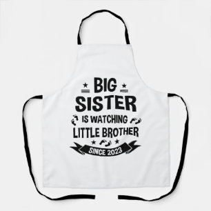 De kleine zus zorgt voor de grote broer in schort