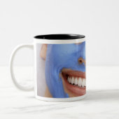 Jonge vrouw die een gelaatsmasker draagt, glimlach tweekleurige koffiemok (Links)