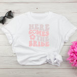 Retro hier komt de bruid vrijgezellenfeest t-shirt<br><div class="desc">Schattigee bruid's vrijgezellenfeest T-shirt met "Here Comes The Bride" in een groot roze en taupe retro 70's stijl lettertype en een illustratie van een madeliefje. Een coördinerend "Here Comes The Party" t-shirt is beschikbaar voor de bachelorette.</div>