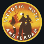 Vintage Travel, Victoria Hotel, Amsterdam, Holland Ronde Sticker<br><div class="desc">illustratie Europese reisposter kunst van het Victoria Hotel,  Amsterdam,  Nederland met een man en een vrouw in klederdracht en het dragen van klompen. Een zeilboot op de oceaan,  een gele zonsondergang en een traditionele windmolen liggen op de achtergrond achter een silhouet van een gelukkig stel.</div>