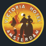 Vintage Travel, Victoria Hotel, Amsterdam, Holland Ronde Sticker<br><div class="desc">illustratie Europese reisposter kunst van het Victoria Hotel,  Amsterdam,  Nederland met een man en een vrouw in klederdracht en het dragen van klompen. Een zeilboot op de oceaan,  een gele zonsondergang en een traditionele windmolen liggen op de achtergrond achter een silhouet van een gelukkig stel.</div>